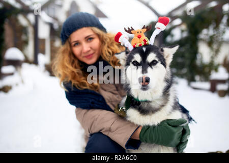 Attraktive authentische kaukasische Frau umarmt Lustig malamute Hund santa tragen liebe Weihnachten Geweih. Curly lächelnd weibliche Spaß mit huskie Welpen im neuen Jahr. Pet ist das beste Geschenk für Feiertage. Stockfoto