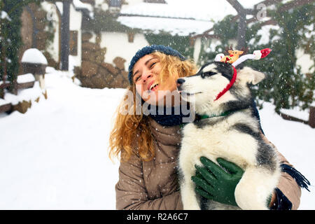 Attraktive kaukasische Frau umarmt Lustig malamute Hund santa tragen liebe Weihnachten Geweih. Curly lächelnd weibliche Spaß mit huskie Welpen im neuen Jahr. Pet ist das beste Geschenk. Doggy licking Girl's Gesicht. Stockfoto