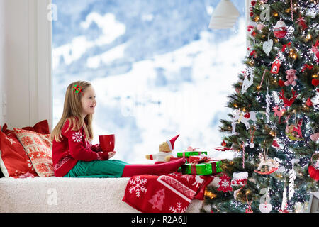 Kind trinken heiße Schokolade am Weihnachtsbaum zu Hause. Kid öffnen Geschenke und Geschenke. Kleines Mädchen in Santa kleid und hut mit heißem Kakao in Rot Stockfoto