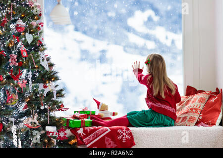 Kind trinken heiße Schokolade am Weihnachtsbaum zu Hause. Kid öffnen Geschenke und Geschenke. Kleines Mädchen in Santa kleid und hut mit heißem Kakao in Rot Stockfoto