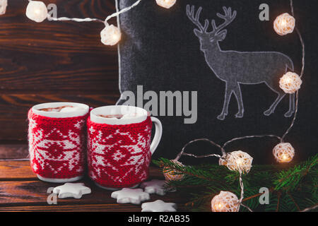 Heiße Schokolade mit Marshmallows und Keksen auf Weihnachtsschmuck Hintergrund. Kissen mit einem Reh, Garland und Tanne gemütliche Atmosphäre schaffen. Stockfoto