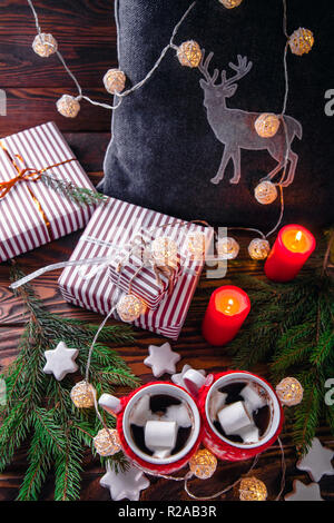 Heiße Schokolade mit Marshmallows und Keksen auf Weihnachtsschmuck Hintergrund. Kissen mit einem Reh, Garland und Tanne gemütliche Atmosphäre schaffen. Stockfoto