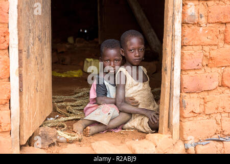 Zwei ugandische Kinder in zerlumpter Kleidung in einer Umarmung auf einem unbefestigten Boden an der Tür eines Hauses sitzen. Stockfoto