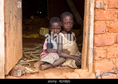 Zwei ugandische Kinder in zerlumpter Kleidung in einer Umarmung auf einem unbefestigten Boden an der Tür eines Hauses sitzen. Stockfoto