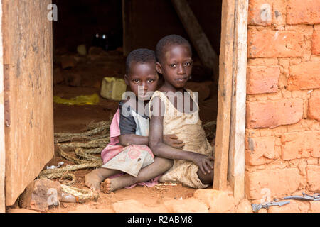 Zwei ugandische Kinder in zerlumpter Kleidung in einer Umarmung auf einem unbefestigten Boden an der Tür eines Hauses sitzen. Stockfoto