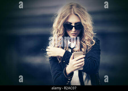 Blonf business Frau mit smart phone in city street Stilvolle Mode Modell mit Langen lockigen Haare mit schwarzen Jacke und Sonnenbrille Stockfoto