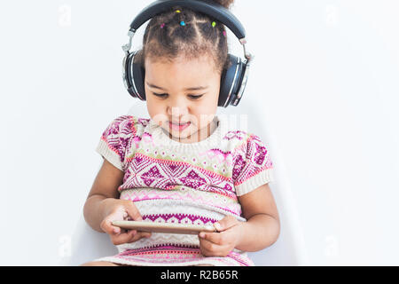 Süße kleine Mädchen in einem bunten Kleid, hört Musik mit Kopfhörern und verwendet ein Smartphone auf weißem Hintergrund Stockfoto