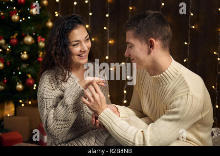 Der Mann gibt dem Mädchen einen Verlobungsring, Paar in Weihnachtsbeleuchtung und Dekoration in Weiß, Tanne auf dunklem Hintergrund gekleidet, winter Stockfoto