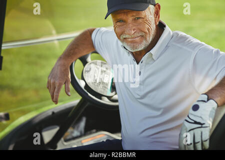 Lächelnd älterer Mann sitzt in einem Warenkorb während einer Runde Golf genießen Sie an einem sonnigen Tag Stockfoto