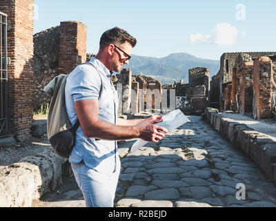 Schöner Mann in Sonnenbrille auf eine Landkarte mit Sehenswürdigkeiten vor dem Hintergrund der historischen Ruinen, blauer Himmel und die Strahlen der untergehenden Sonne suchen. Italien, Po Stockfoto