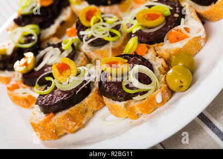 Traditionelle spanische Blutwurst serviert auf frischem Baguette mit grünen Oliven und in Scheiben geschnittenen Lauch Stockfoto