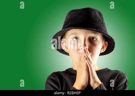 Junge nachdenkliche Junge tief in Gedanken versunken, hält die Handflächen zusammen auf Mund, grüne Augen, trägt schwarze Hut grün studio Wand mit kostenlosen Speicherplatz für die Stockfoto