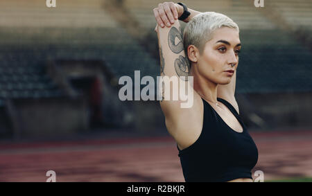 Seitenansicht Porträt einer Läuferin zu tun Stretching Übungen in einem Leichtathletik Stadion stehen. Frau in Fitness Kleidung ihre Arme stretching s Stockfoto