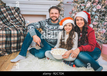 Bild von Familie sitzt auf dem Boden und lächelnd vor der Kamera. Junge Mann hat die Fernbedienung in der Hand. Mädchen hält Schüssel Popcorn. Sie lehnt sich an die Mutter. Stockfoto