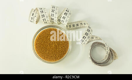 Brauner Zucker in kleinen transparenten Glas bawl und eine gewellte Maßband in Zentimeter daneben vor einem weißen Hintergrund Stockfoto