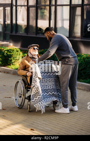 Junge afrikanische amerikanische cuidador für ältere behinderte Menschen im Rollstuhl mit Plaid auf der Straße Stockfoto