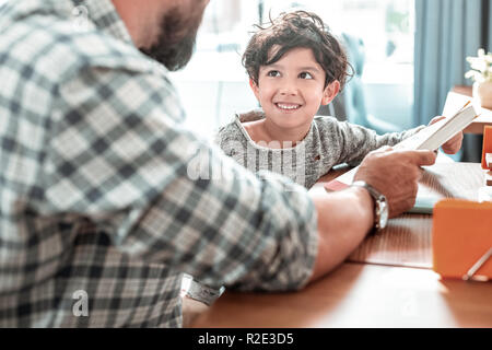 Süß lächelnd dark-eyed Sohn bei seinem Vater tragen squared Shirt suchen Stockfoto