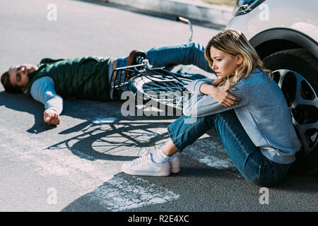 Hohe Betrachtungswinkel und der jungen Frau in der Nähe von Auto und Opfer eines Autounfalls liegen auf der Straße sitzen Stockfoto