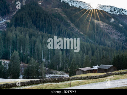 Berg und Tal Landschaft mit Wald und schneebedeckte Berggipfel und einer alten Felswand mit Hütten und chalets hinter der Sonne und Stockfoto