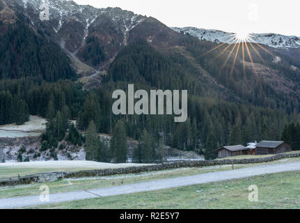 Berg und Tal Landschaft mit Wald und schneebedeckte Berggipfel und einer alten Felswand mit Hütten und chalets hinter der Sonne und Stockfoto