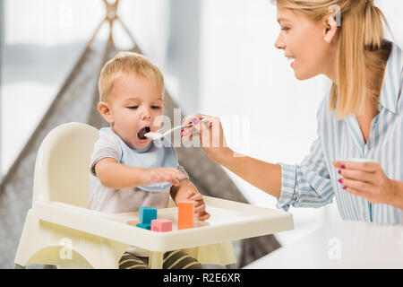 Mutter Fütterung Sohn im Hochstuhl mit Babynahrung Stockfoto