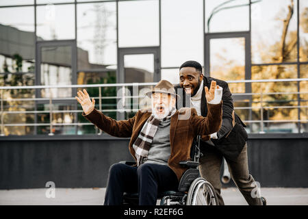 Gerne ältere behinderte Menschen im Rollstuhl und African American man Spaß beim Reiten durch die Straße Stockfoto