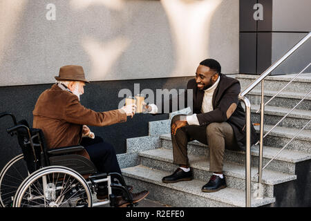 Ältere behinderte Menschen im Rollstuhl und afrikanische amerikanische Mann Kaffee zusammen trinken und klirren Pappbecher auf der Straße Stockfoto