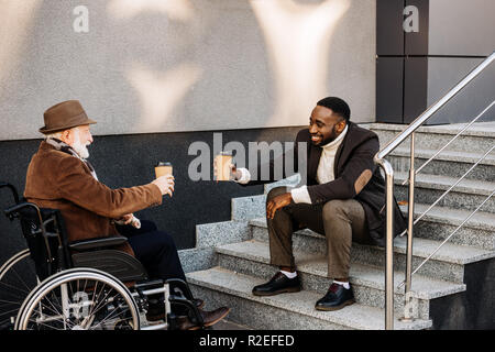 Gerne ältere behinderte Menschen im Rollstuhl und afrikanische amerikanische Mann Kaffee zusammen trinken und klirren Pappbecher auf der Straße Stockfoto