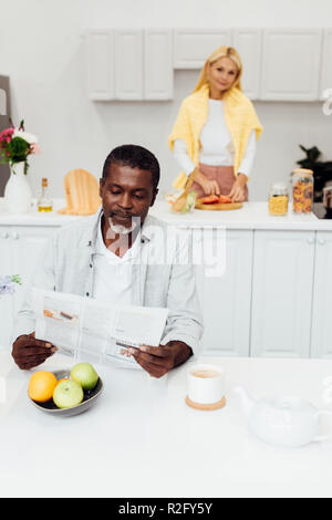 Afrikanische amerikanische Mann lesen Zeitung, während Frau kochen Frühstück Stockfoto