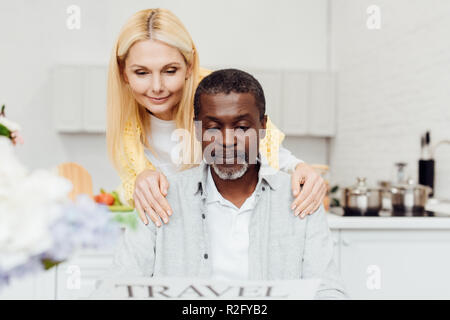 Reifen lächelnde Frau umarmt african american man während Reisen Zeitung an Küche lesen Stockfoto