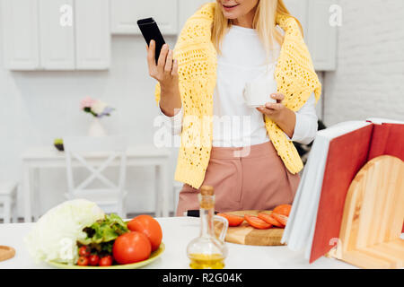 7/8-Ansicht der blonden Frau mit Smartphone beim Kochen Abendessen Stockfoto