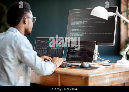 Junge afrikanische Mann Programmierer schreiben Programmcode am Arbeitsplatz sitzen mit drei Monitoren im Büro. Bild auf dem Bildschirm Stockfoto