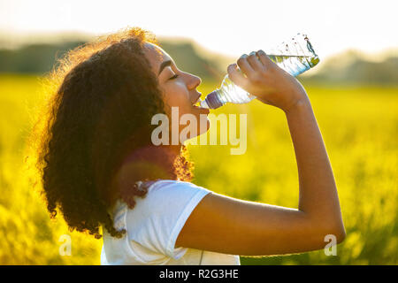 Outdoor Portrait schöne glückliche Mischlinge afroamerikanische Mädchen Teenager weibliche junge Frau Trinkwasser aus einer Flasche in einem Feld von gelben flo Stockfoto