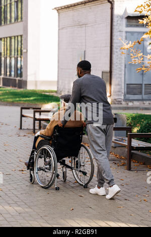 Ansicht der Rückseite des älteren Behinderten Mann im Rollstuhl und Krankenschwester Reiten auf der Straße Stockfoto