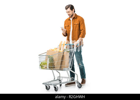 Glücklich aussehenden Mann, der Einkaufswagen mit den Produkten in Papiertüten und Sie ihr Smartphone isoliert auf weißem Stockfoto