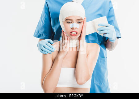 Chirurg in Handschuhe bis taping Frau Gesicht mit Bandage nach plastische Chirurgie isoliert auf weißem Stockfoto