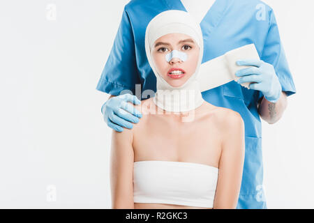Chirurg in Handschuhe bis taping Frau Gesicht mit Bandage nach plastische Chirurgie isoliert auf weißem Stockfoto