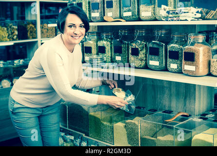 Reifen lächelt froh, Frau neben Gläsern mit getrocknete Getreide und Kommissionierung in organischen Store ausgewählt Stockfoto