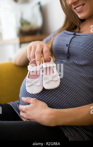 Schwangere Frau Sittingh auf dem Sofa zu Hause Holding Baby Schuhe Stockfoto