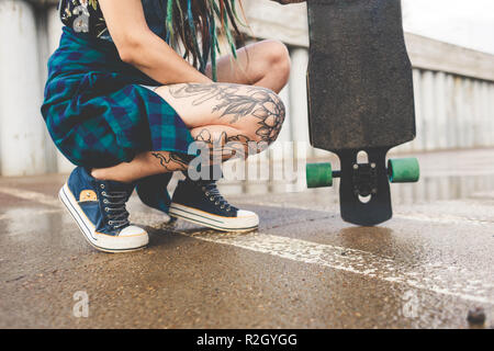 Junges Mädchen mit Tattoos und Dreadlocks sitzt mit einem Longboard auf dem Hintergrund der konkreten Struktur Stockfoto