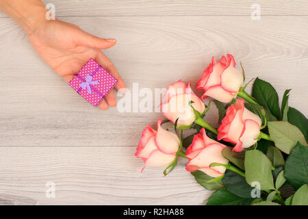 Woman's Hände halten ein Geschenk Box über graues Holz- Hintergrund mit wunderschönen Rosen. Konzept der ein Geschenk an Feiertagen. Ansicht von oben. Stockfoto