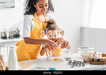 Lächelnd afrikanische amerikanische Mutter, Tochter brechen Ei für die Zubereitung von Dessert in der Küche Stockfoto