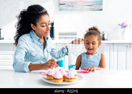 Afrikanische amerikanische Mutter und Tochter mit Tiaras am Tisch sitzen mit Muffins in der Küche Stockfoto