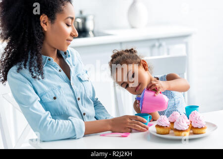 Afrikanische amerikanische Tochter und Mutter spielt mit Plastik Geschirr in der Küche, Kid gießen Kaffee Stockfoto