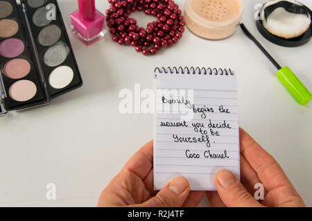Frau Hand hält Coco Chanel Zitate auf einen Block Notiz geschrieben, pearl Zubehör und bis auf weißem Hintergrund machen, inspiration Satz "Schönheit beginnt die Stockfoto