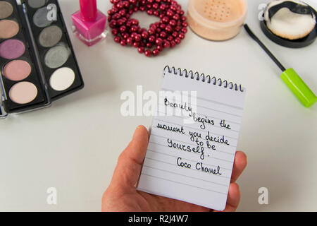 Frau Hand hält Coco Chanel Zitate auf einen Block Notiz geschrieben, pearl Zubehör und bis auf weißem Hintergrund machen, inspiration Satz "Schönheit beginnt die Stockfoto