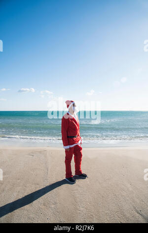 Junger Mann, der eine Santa Anzug stehen auf dem Strand mit Blick auf die Sonne mit dem Meer im Hintergrund Stockfoto