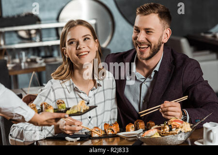 Lächelndes Paar essen Sushi Rollen während der Kellner bringt neue Bestellung im Restaurant Stockfoto