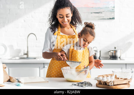 Lächelnd afrikanischen amerikanischen Mutter und Tochter Teig vorbereiten und Sieben Mehl in der Küche Stockfoto