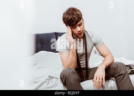 Hübscher junger Geschäftsmann mit Riemen über Hals sitzt auf dem Bett und mit Kopfschmerzen zu Hause Stockfoto